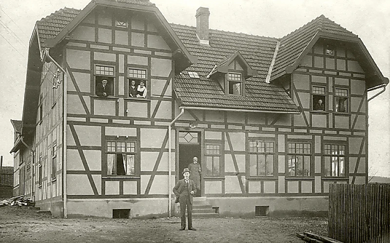Historische Schwarzweißaufnahme eines großen Fachwerkhauses mit Ziegeldach; mehrere Personen stehen an Fenstern und vor dem Hauseingang.